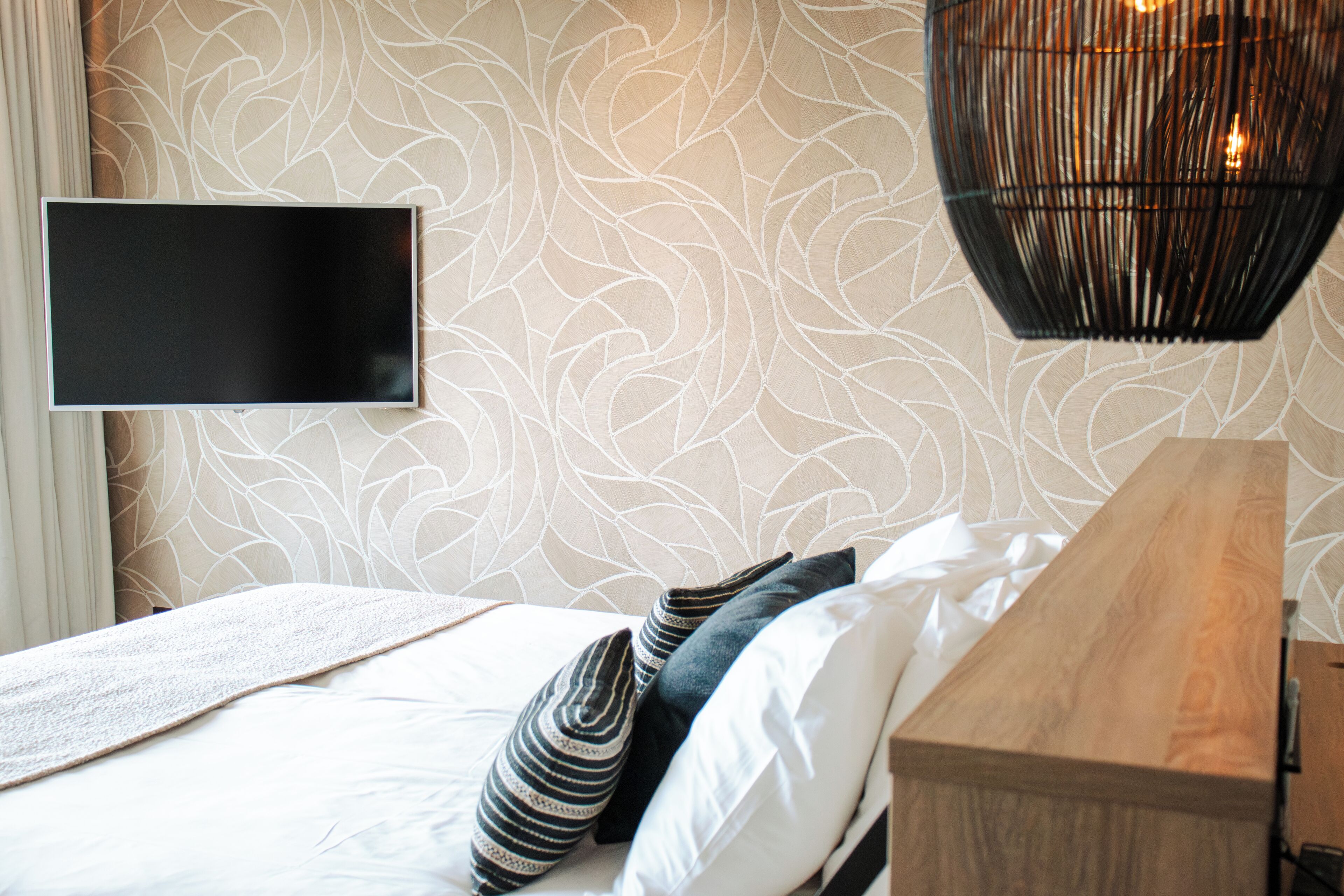 A modern bedroom with a patterned wallpaper, a flat-screen TV mounted on the wall, a wooden bedside table, and a bed with white and striped pillows.