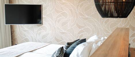A modern bedroom with a patterned wallpaper, a flat-screen TV mounted on the wall, a wooden bedside table, and a bed with white and striped pillows.
