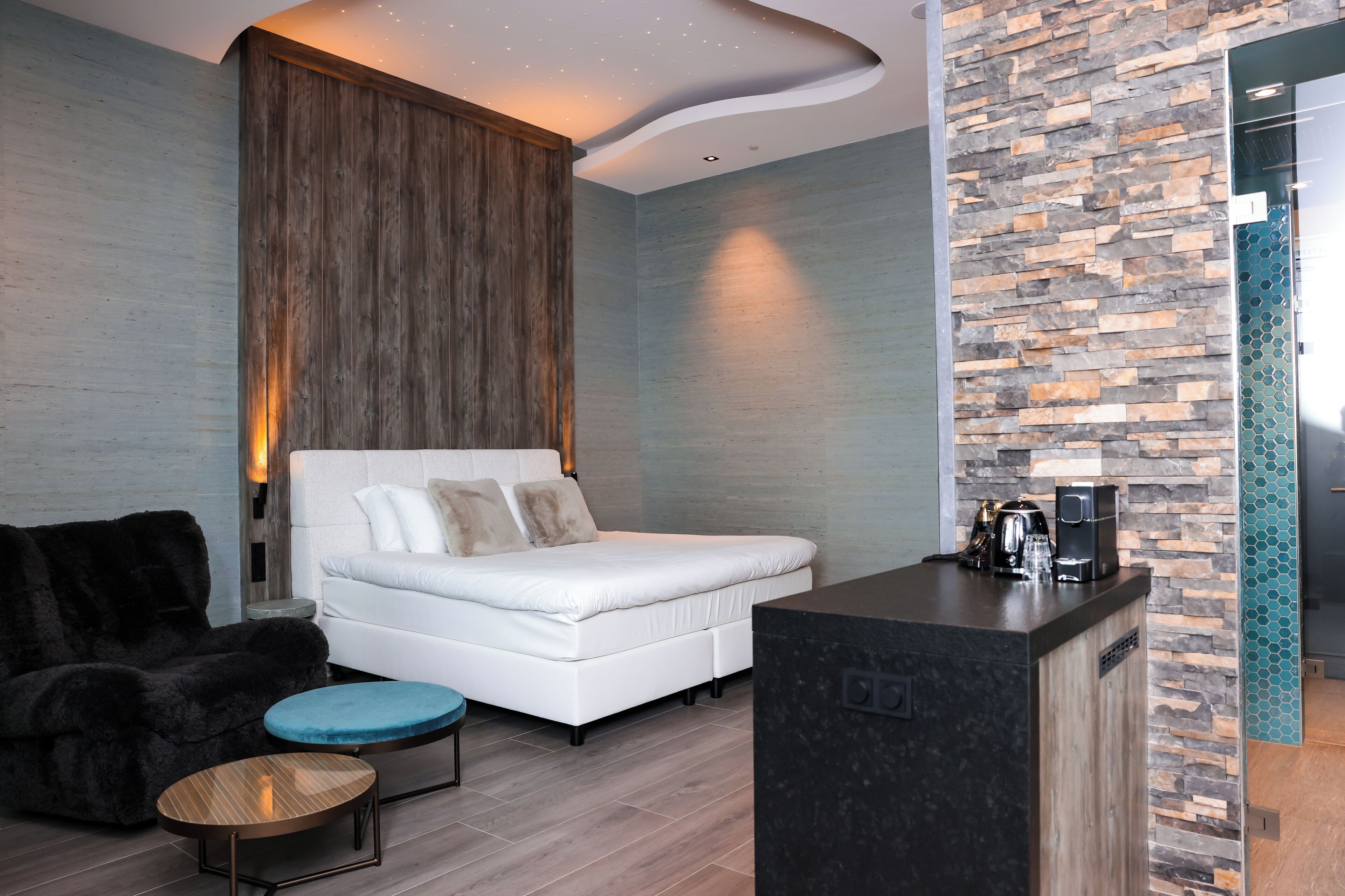 Una habitación de hotel moderna con una cama, una zona de estar con un sillón y reposapiés, una mesita y una kitchenette equipada para café.