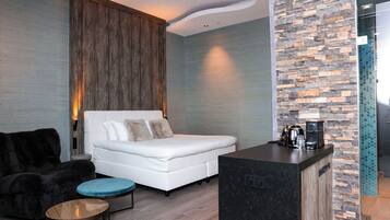 Una habitación de hotel moderna con una cama, una zona de estar con un sillón y reposapiés, una mesita y una kitchenette equipada para café.
