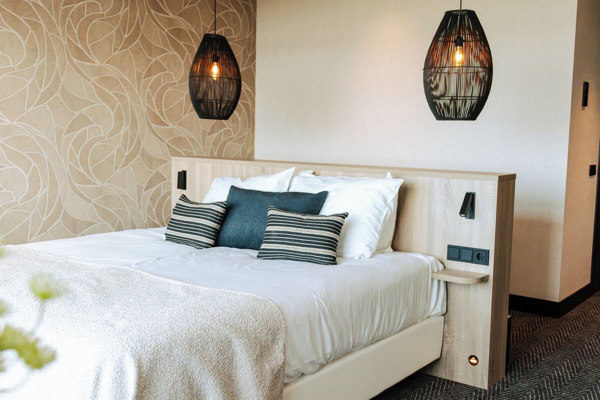 A neatly made bed with white bedding and multiple pillows, a wooden headboard, and two wall-mounted pendant lights.