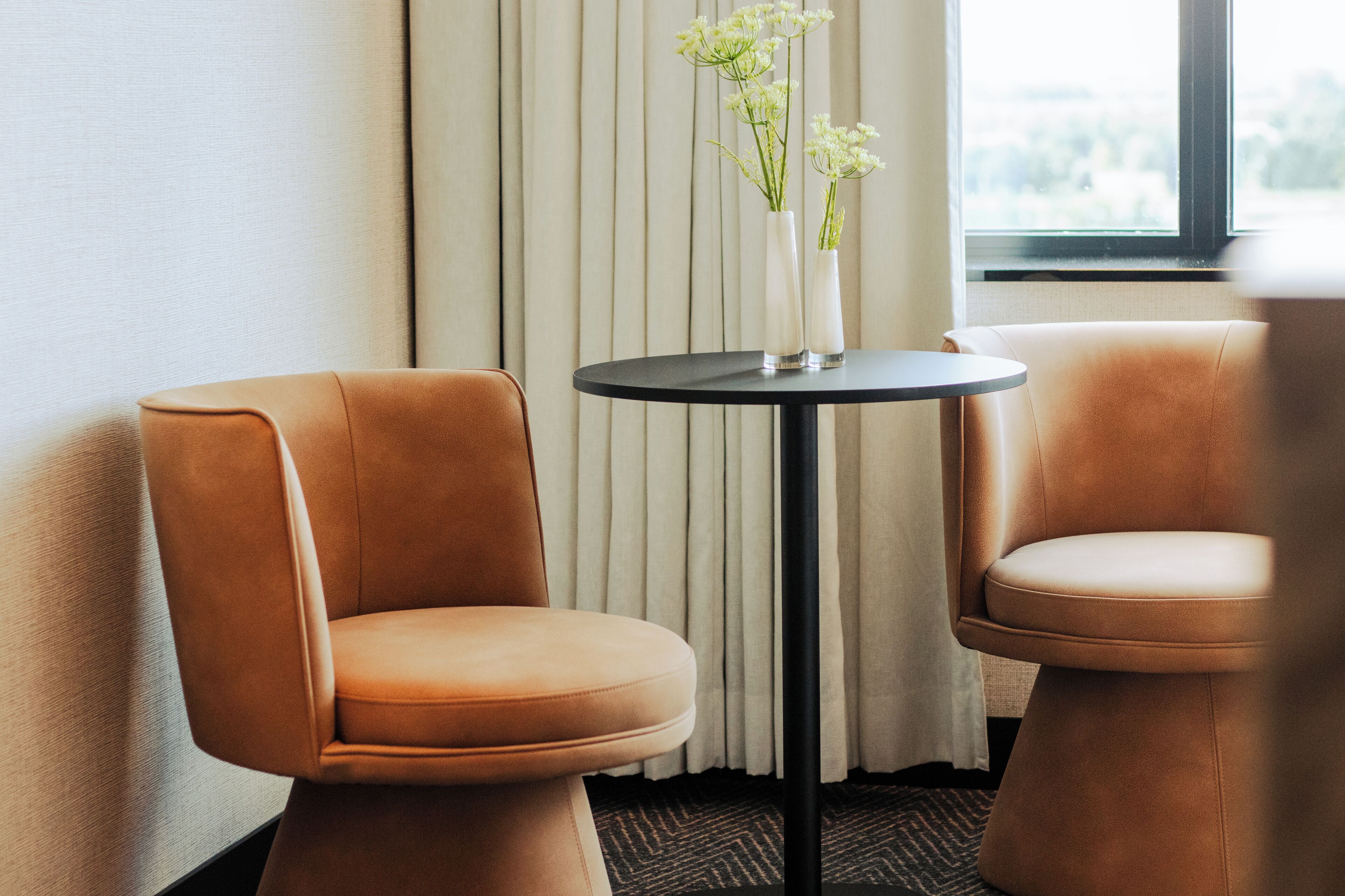 Two brown upholstered chairs with a small round table between them, a white vase with flowers on the table, and a window with curtains in the background.