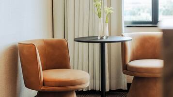 Two brown upholstered chairs with a small round table between them, a white vase with flowers on the table, and a window with curtains in the background.