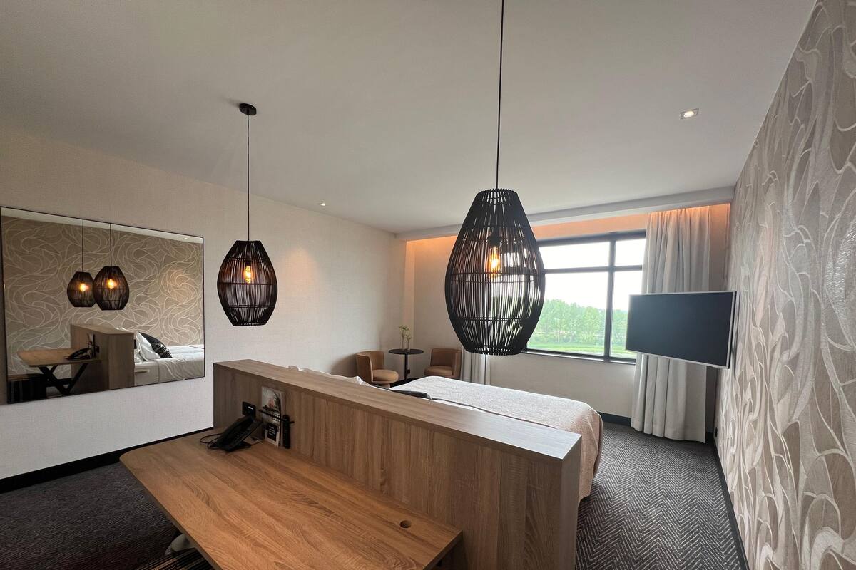 A hotel room with a wooden reception desk, two pendant lights, a large mirror, a flat-screen TV, and a window with curtains.