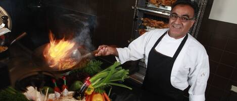 A chef in a kitchen with a grill, vegetables, and cooking utensils.