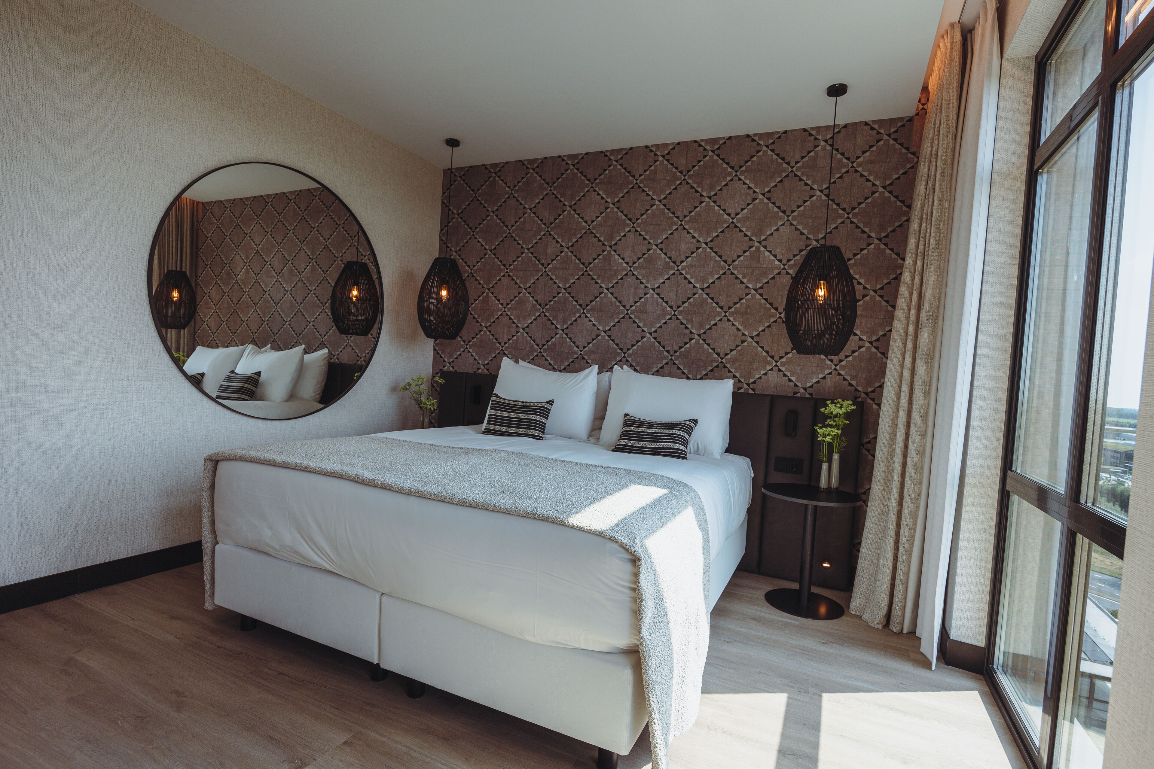 A modern bedroom with a large bed, a round mirror, patterned wallpaper, and pendant lighting.