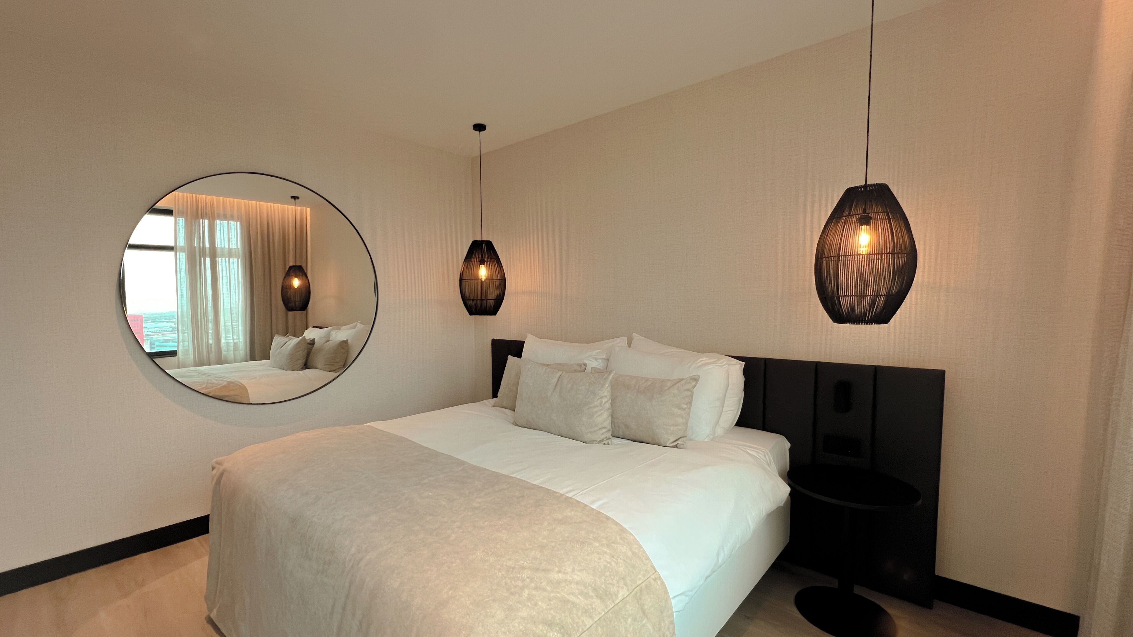 Une chambre d’hôtel avec un grand lit, un miroir rond, deux lampes suspendues et une vue par la fenêtre.