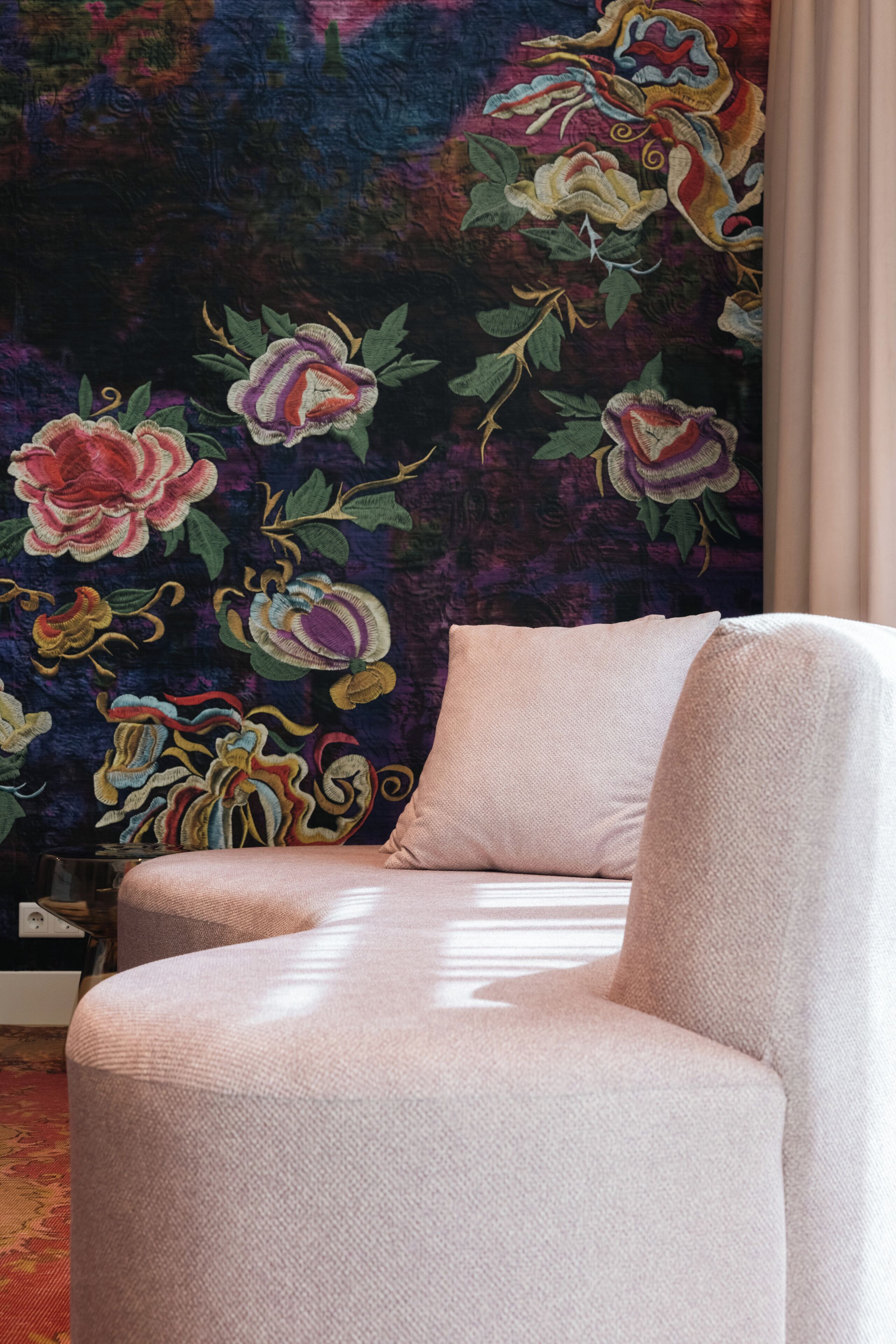 A room with a large floral tapestry on the wall and a light-colored armchair.