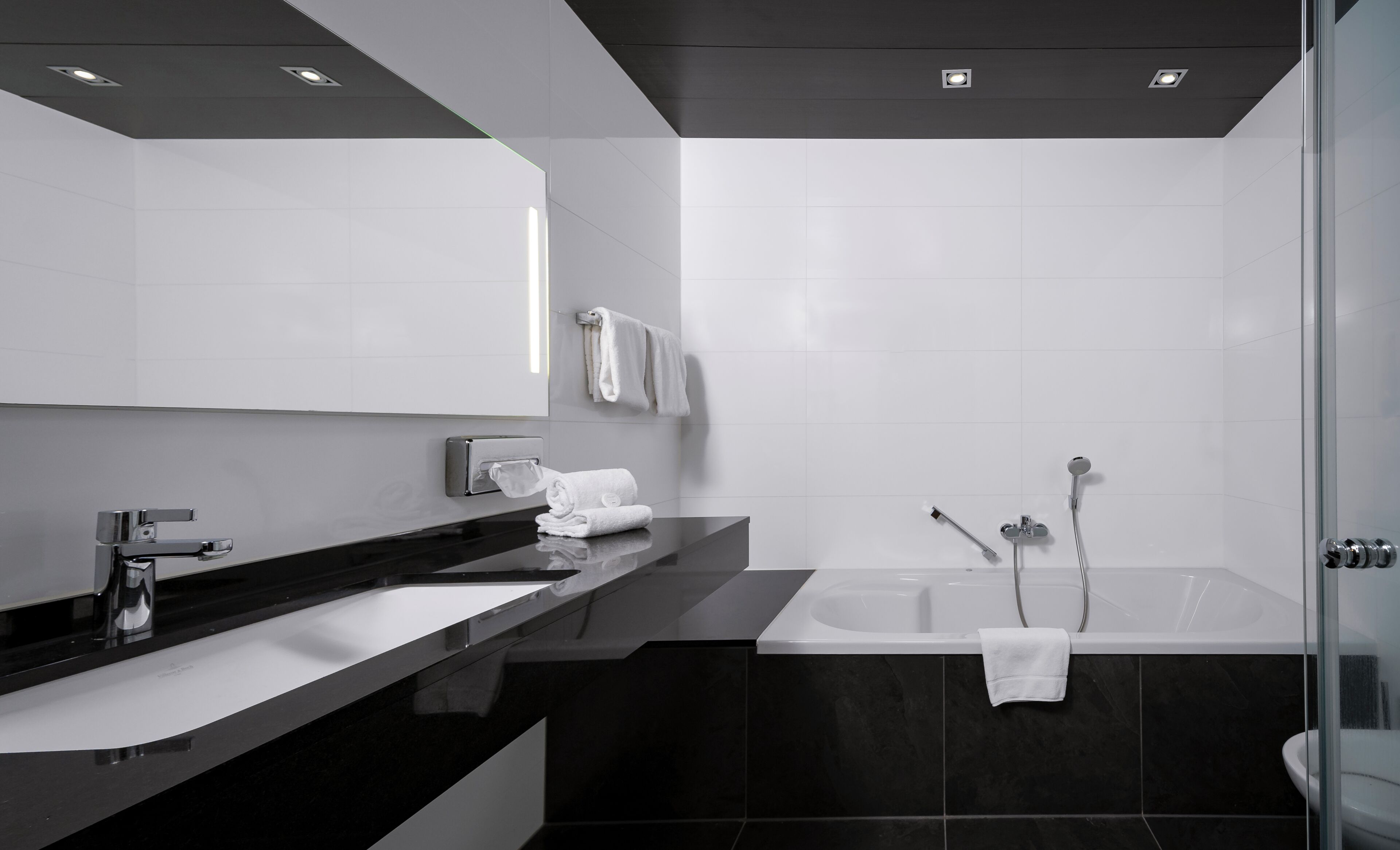 A modern bathroom with a black countertop, white sink, and a large mirror. There is a bathtub with two showerheads and a towel rack with towels.