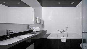 A modern bathroom with a black countertop, white sink, and a large mirror. There is a bathtub with two showerheads and a towel rack with towels.