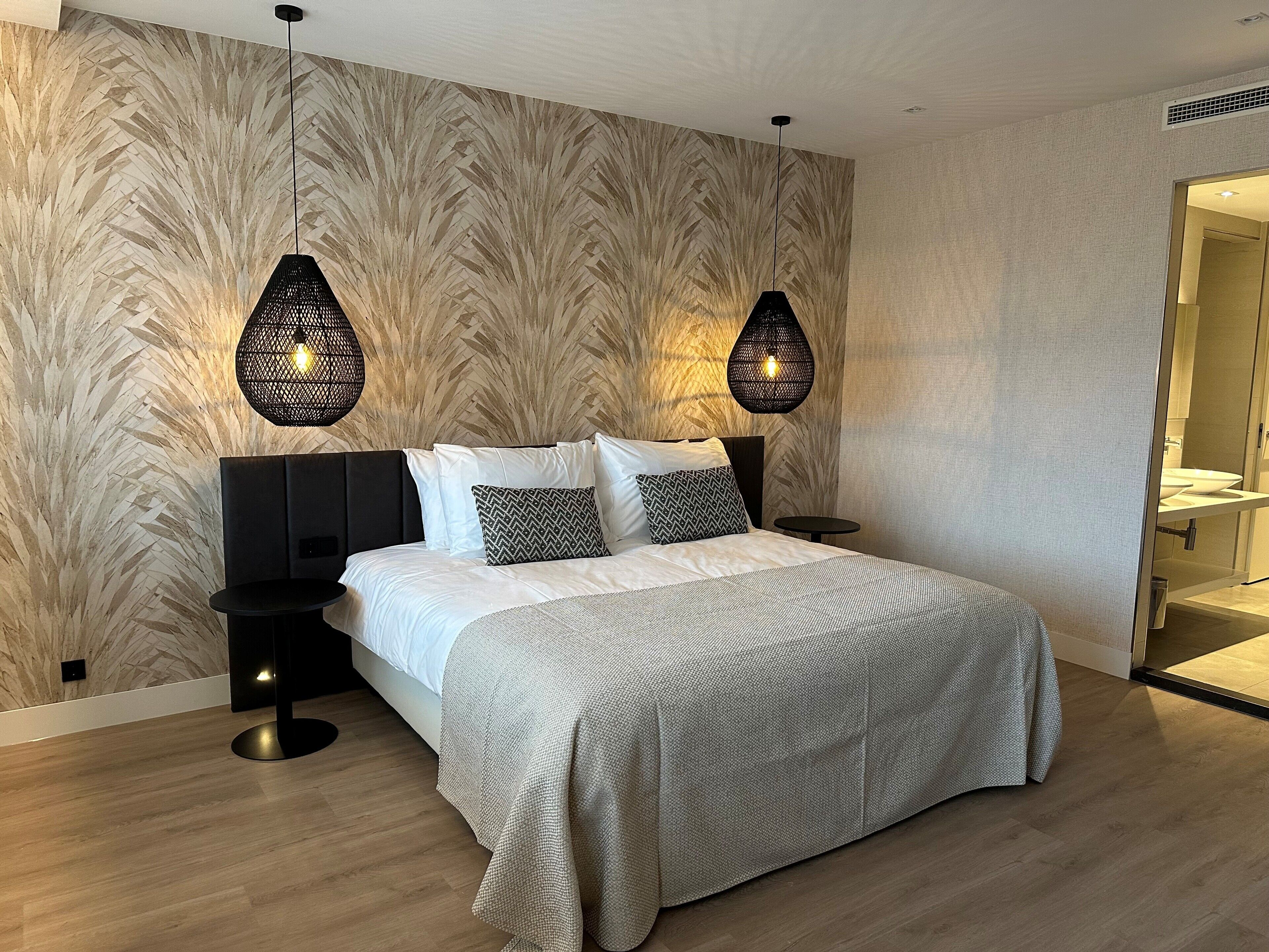 Een moderne slaapkamer met een groot bed, twee nachtkastjes, een ronde tafel en een badkamer zichtbaar door een geopende deur.