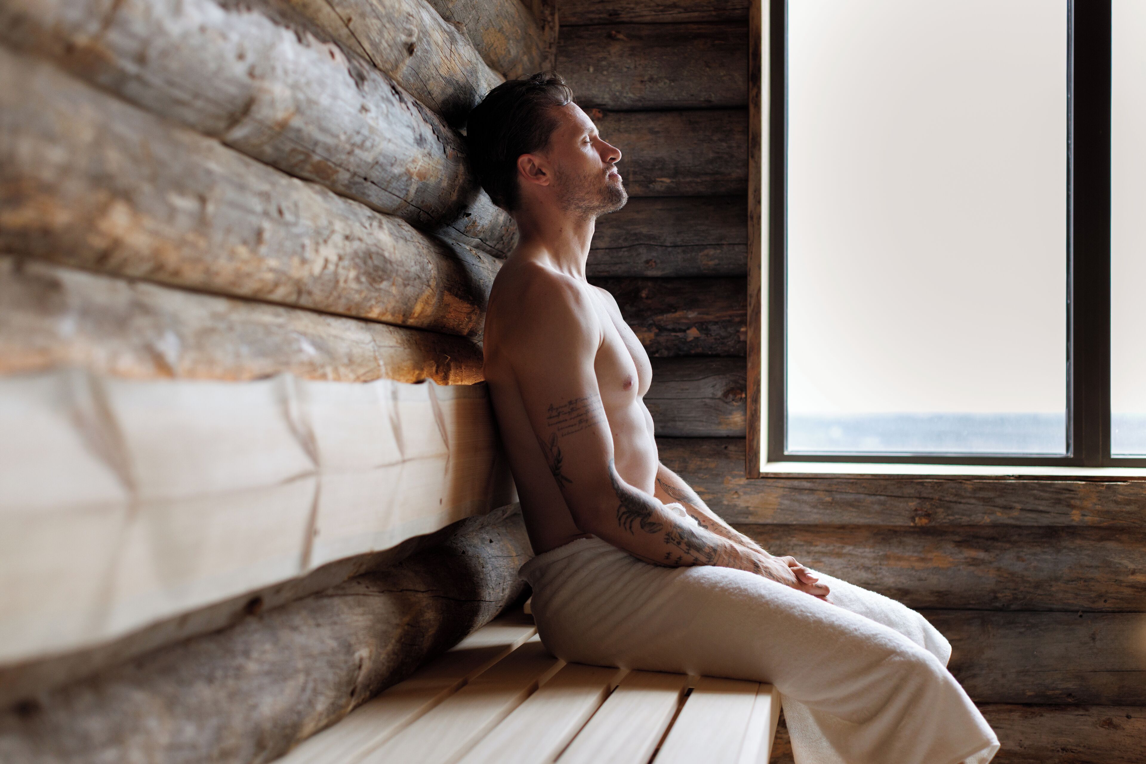 A person sitting on a wooden bench in a log cabin room with a window.