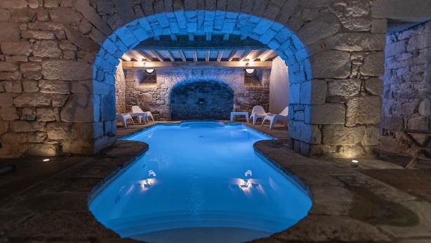 Habitación de paredes de piedra con una piscina central, bancos de piedra y una chimenea de piedra.