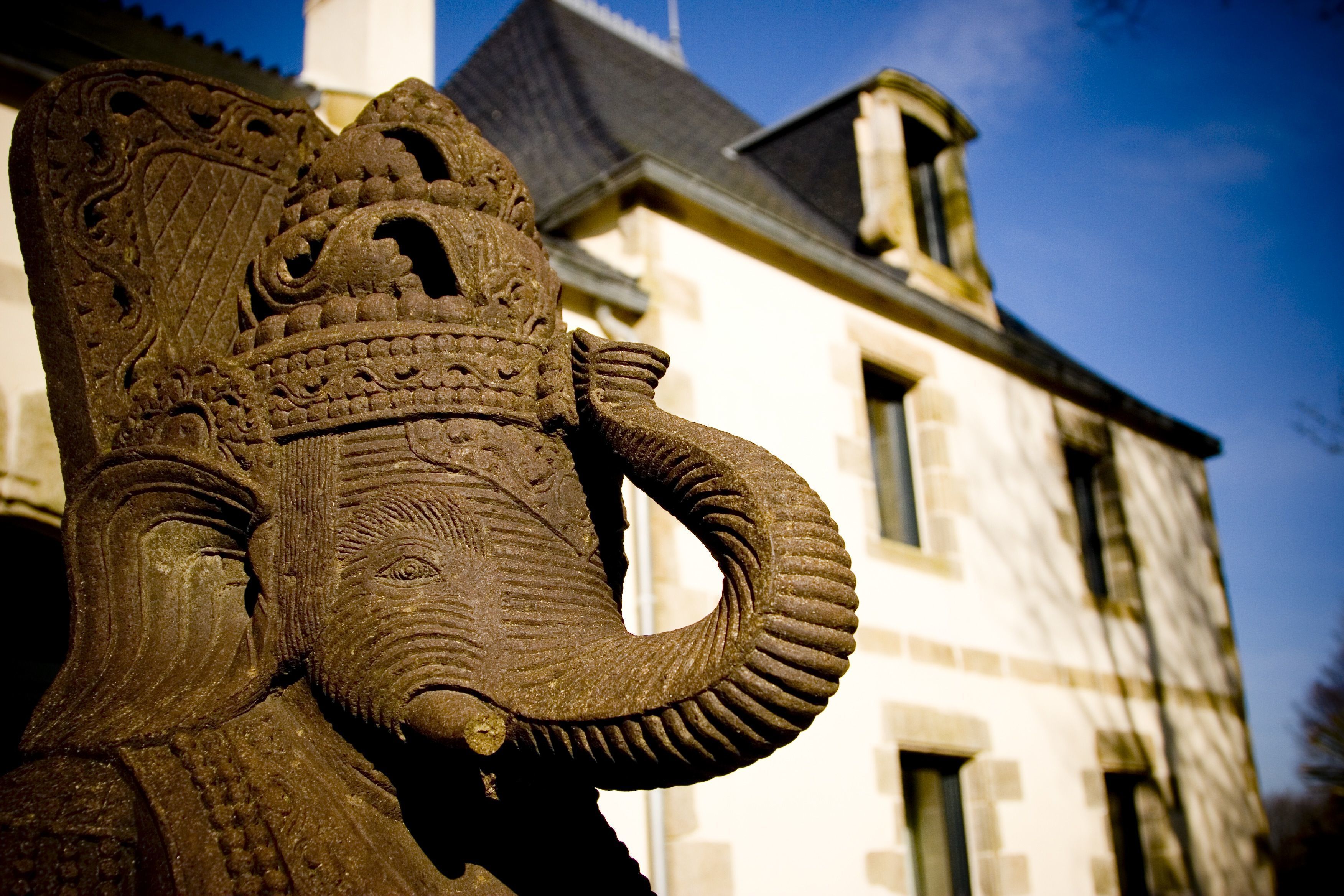 Una escultura de elefante de piedra frente a un edificio con tejado a dos aguas y varias ventanas.