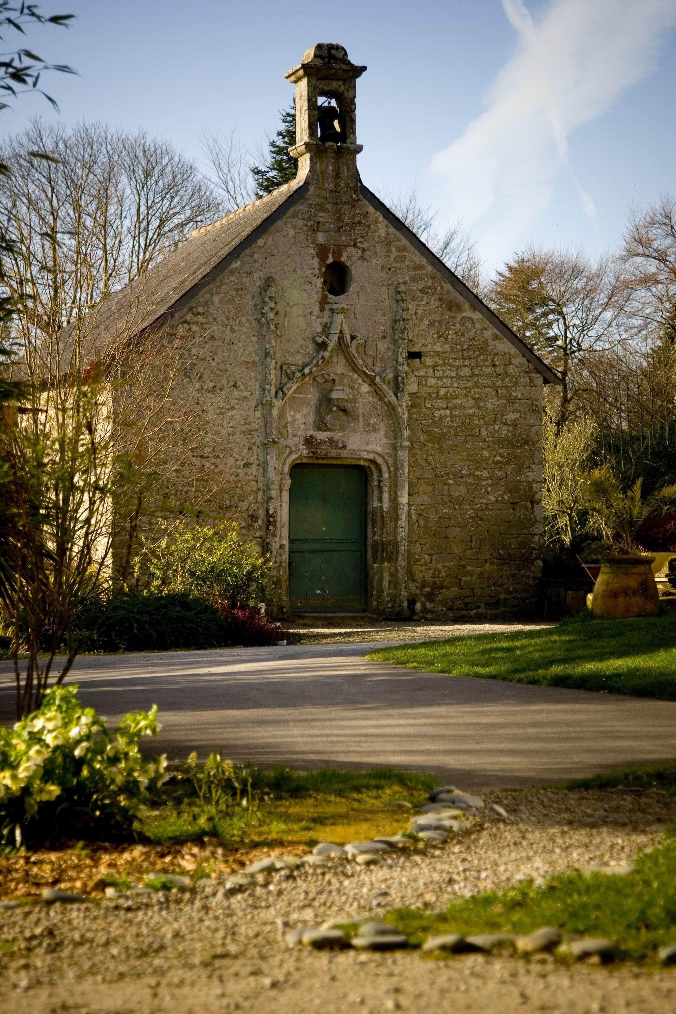 A stone building with a green door, surrounded by greenery and a gravel path.