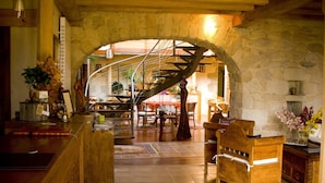 Un interior acogedor con un mostrador de recepción de madera, una escalera de caracol, paredes de piedra y un comedor con sillas y mesas.