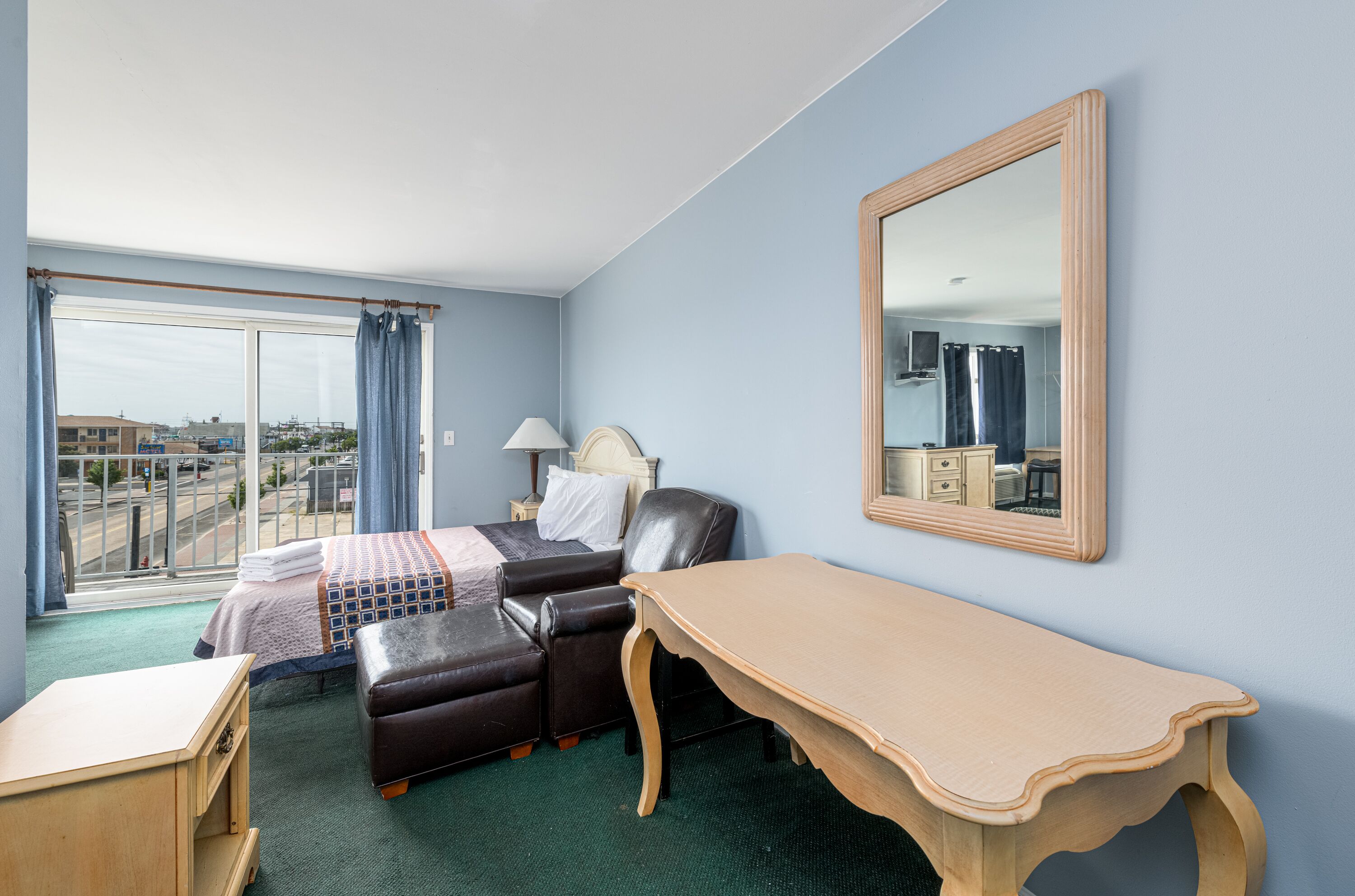 A hotel room with a bed, a desk, a chair, a mirror, and a view of the city through large windows.