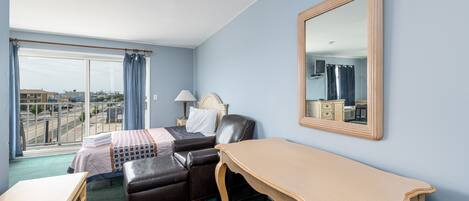 A hotel room with a bed, a desk, a chair, a mirror, and a view of the city through large windows.