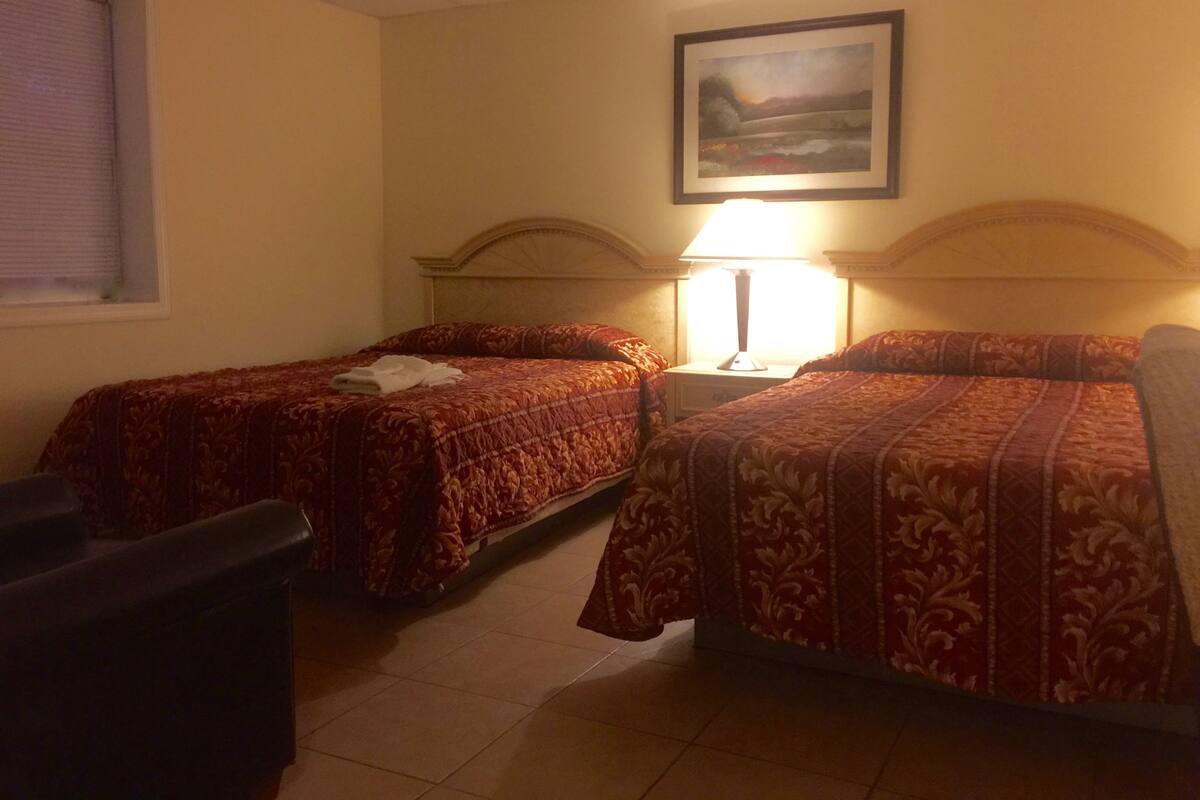 Una habitación de hotel con dos camas individuales, una mesita de noche con lámpara, un cuadro en la pared y una ventana con persianas.