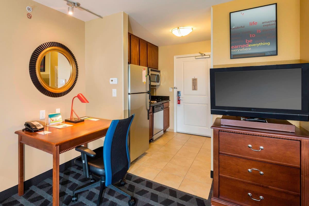 A hotel room with a wooden desk, a chair, a television, a microwave, a refrigerator, a small kitchen area, a mirror, and a wall with motivational words.