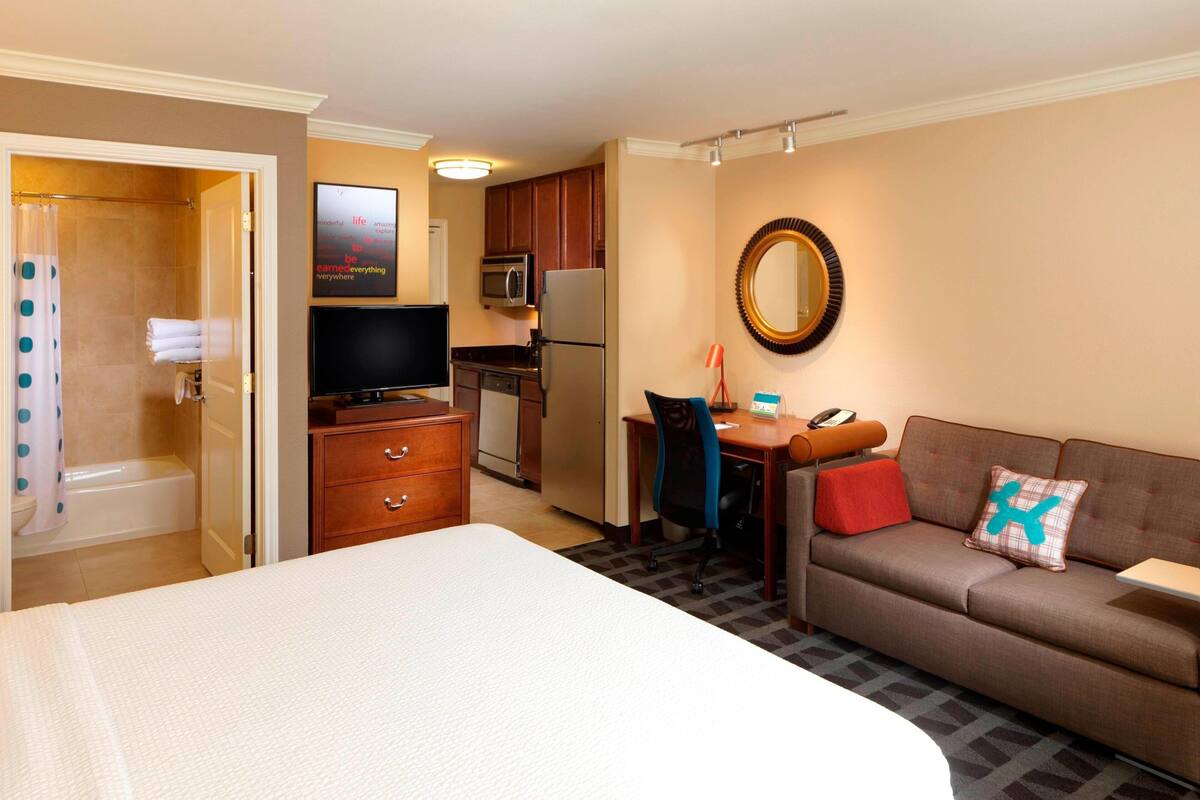 A hotel room with a bed, a sofa, a desk, a TV, a microwave, a refrigerator, and a shower.