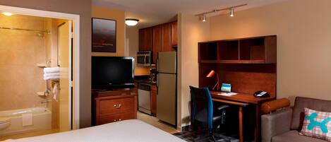 Quarto de hotel com cama, escrivaninha, cadeira, cozinha compacta e área de chuveiro.