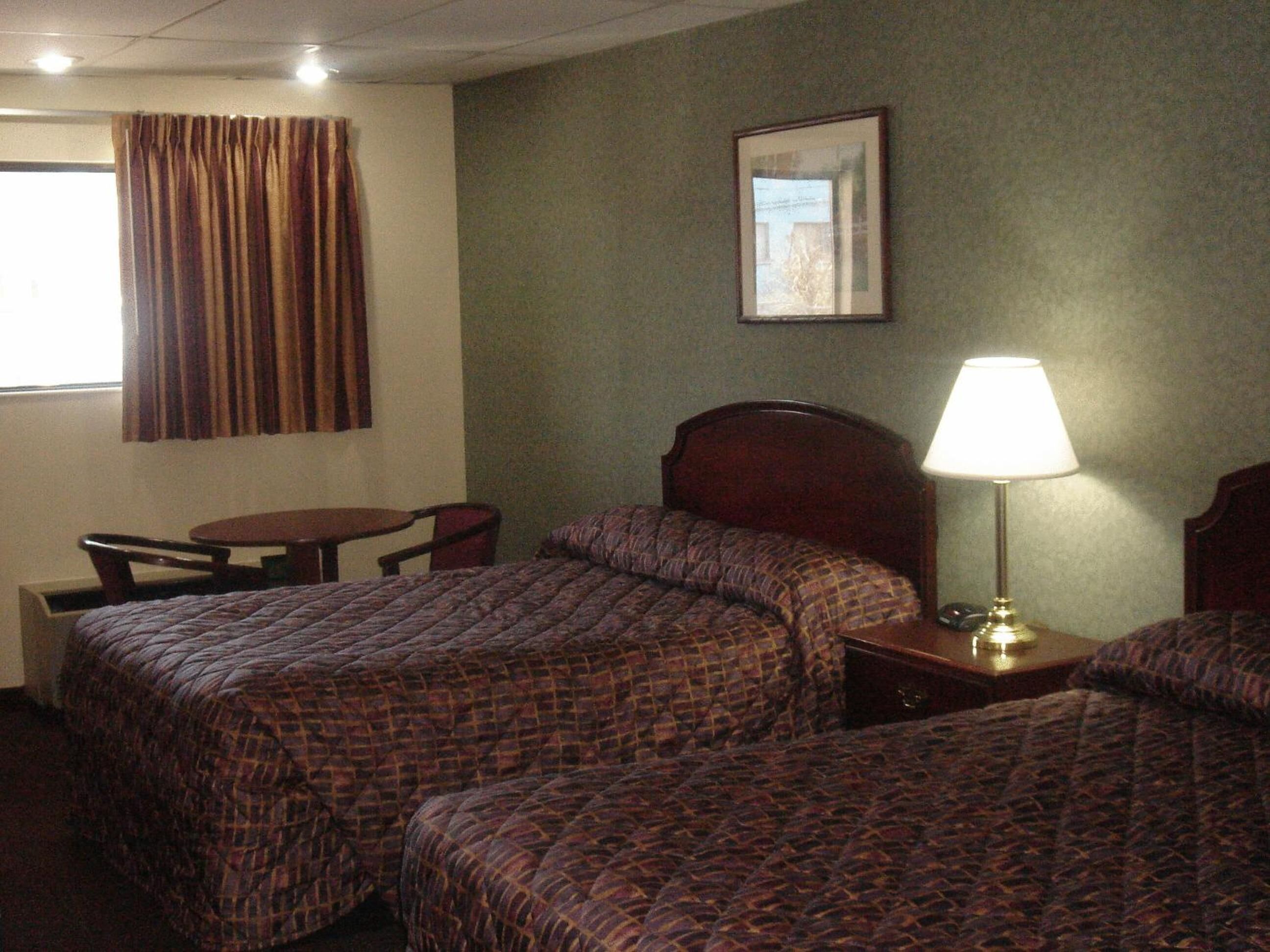 A hotel room with two beds, a small table, a chair, a lamp, and a framed picture on the wall.