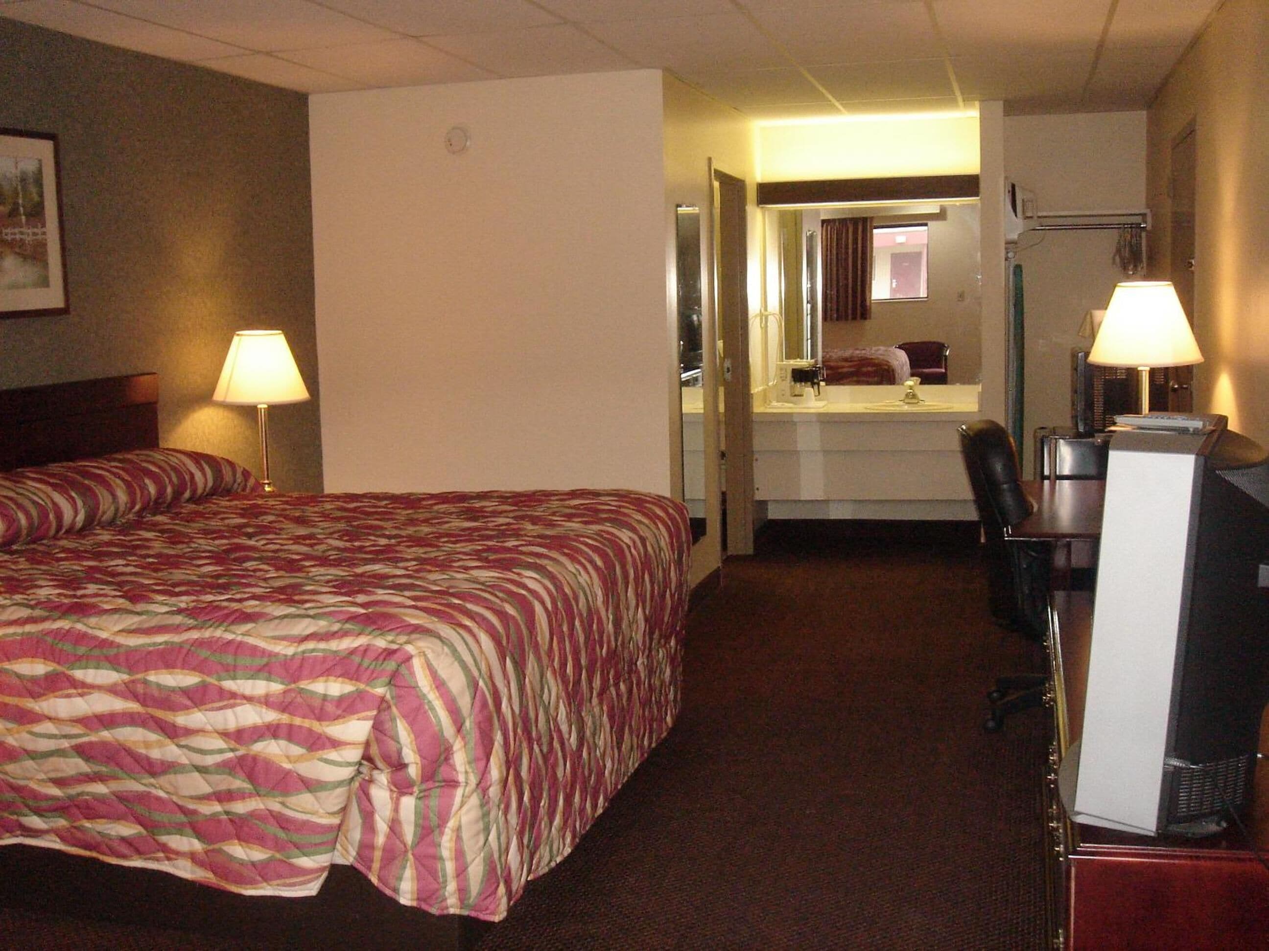 A hotel room with a bed, a desk with a computer, a chair, a desk lamp, and a mirror.