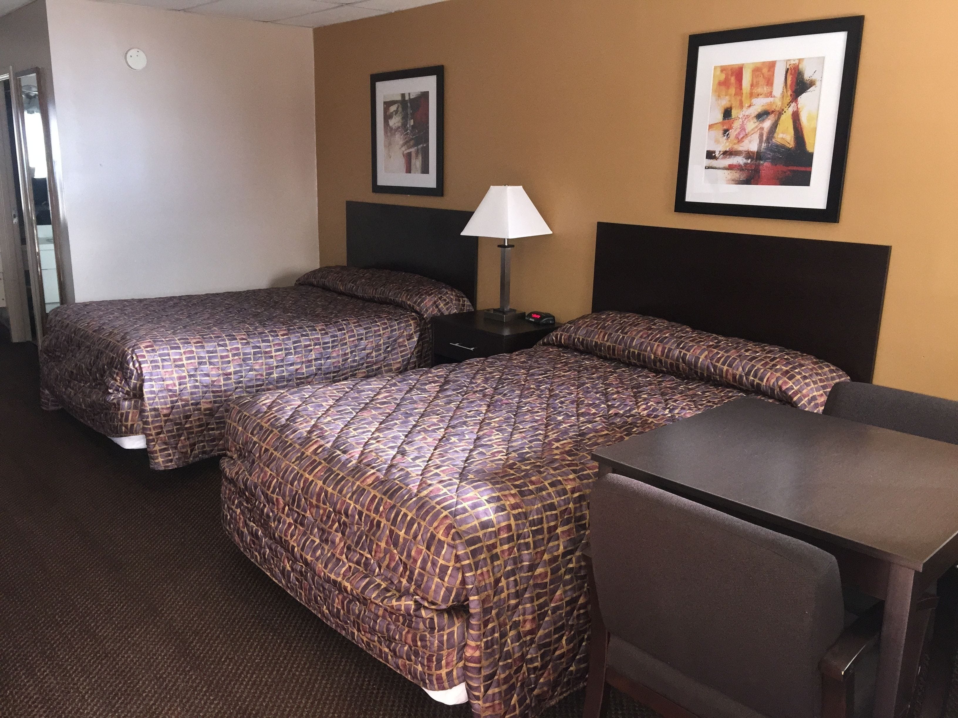 A hotel room with two beds, a desk, a chair, a lamp, and two framed pictures on the wall.