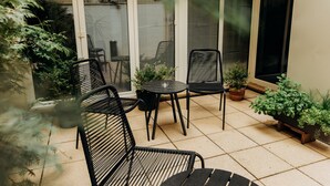 Un patio con una mesa redonda y cuatro sillas, rodeado de plantas en macetas y una puerta de vidrio que da a un balcón.