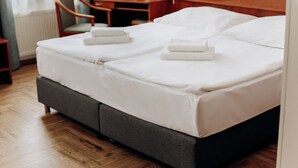Una cama bien hecha con sábanas blancas y dos toallas dobladas, una mesita de noche con una lámpara, y una silla en una habitación de hotel.