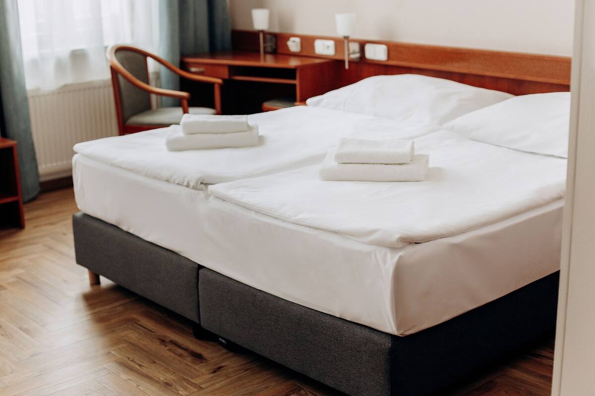 Cama bem arrumada com lençóis brancos e duas toalhas dobradas, mesa de cabeceira de madeira com abajur, e uma cadeira em um quarto de hotel.