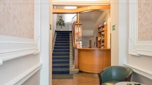 Zona de recepción de un hotel, con un mostrador, una silla verde, una mesita con un cuenco y una escalera que sube a otro piso.