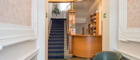 Zona de recepción de un hotel, con un mostrador, una silla verde, una mesita con un cuenco y una escalera que sube a otro piso.