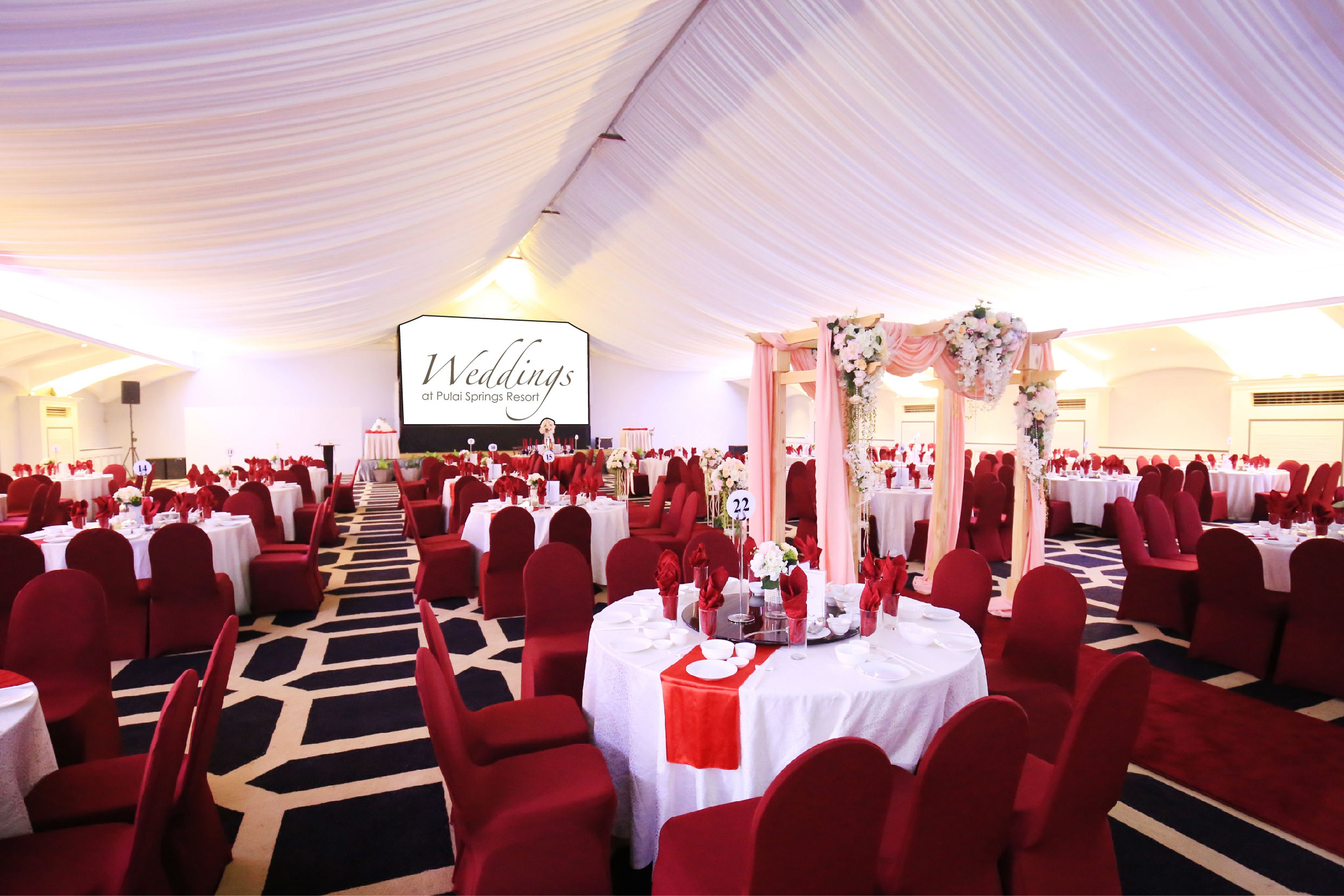 A banquet hall with round tables, red chairs, and a stage with a floral arch.