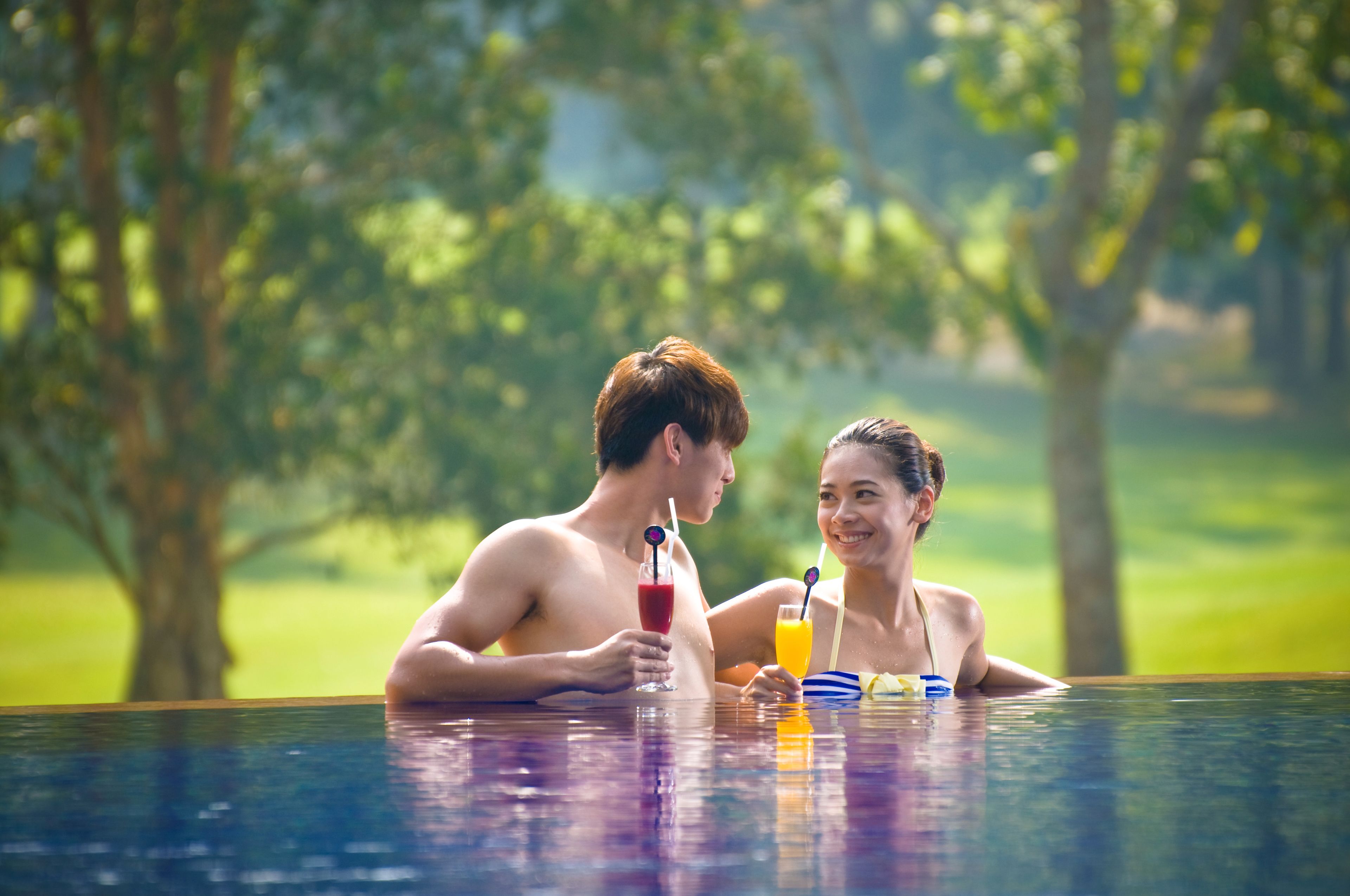 A couple in a swimming pool with drinks, surrounded by greenery.
