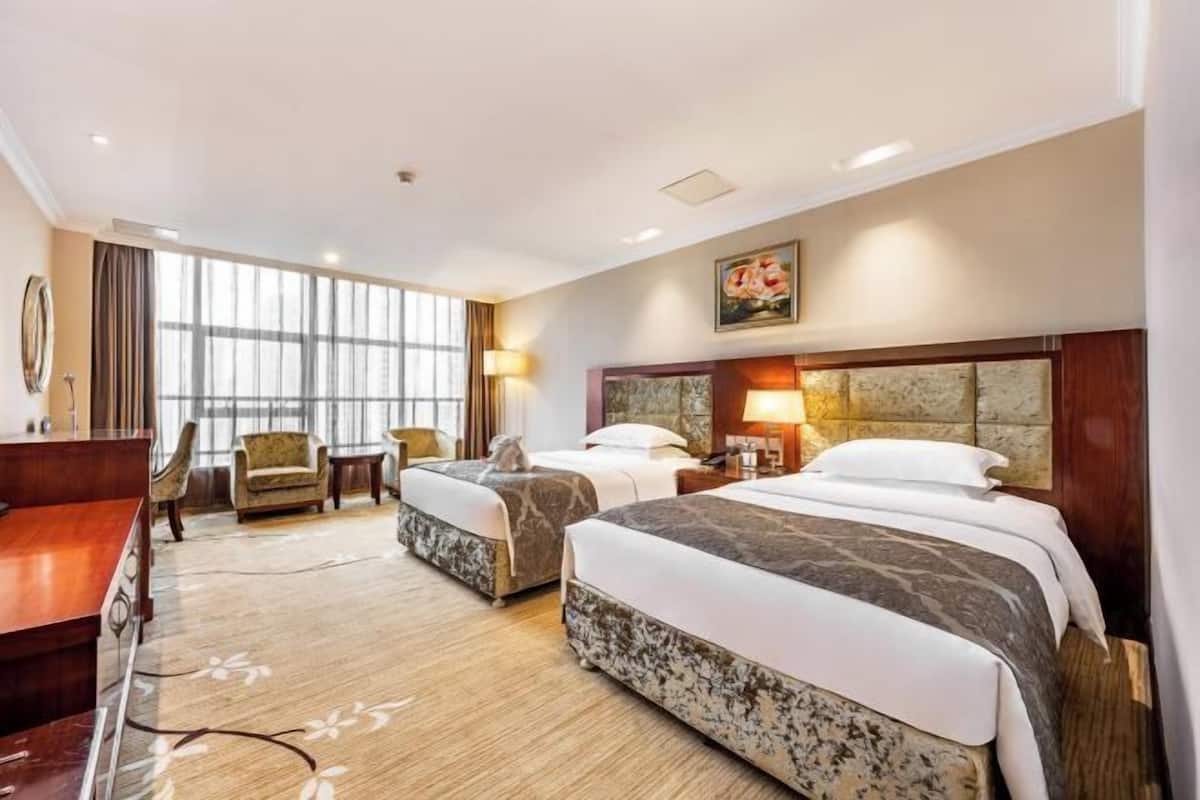 A hotel room with two beds, a desk, a chair, and large windows.