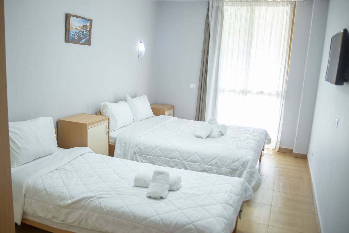 Habitación de hotel con dos camas, una mesita de noche, un televisor montado en la pared y una ventana con cortinas.