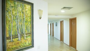 A hallway with a framed painting of trees, a wall-mounted light fixture, and wooden doors. - Hotel Oaz (Golem)