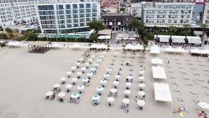 An aerial view of a beach with rows of sun loungers and umbrellas, surrounded by buildings and palm trees. - Hotel Oaz (Golem)