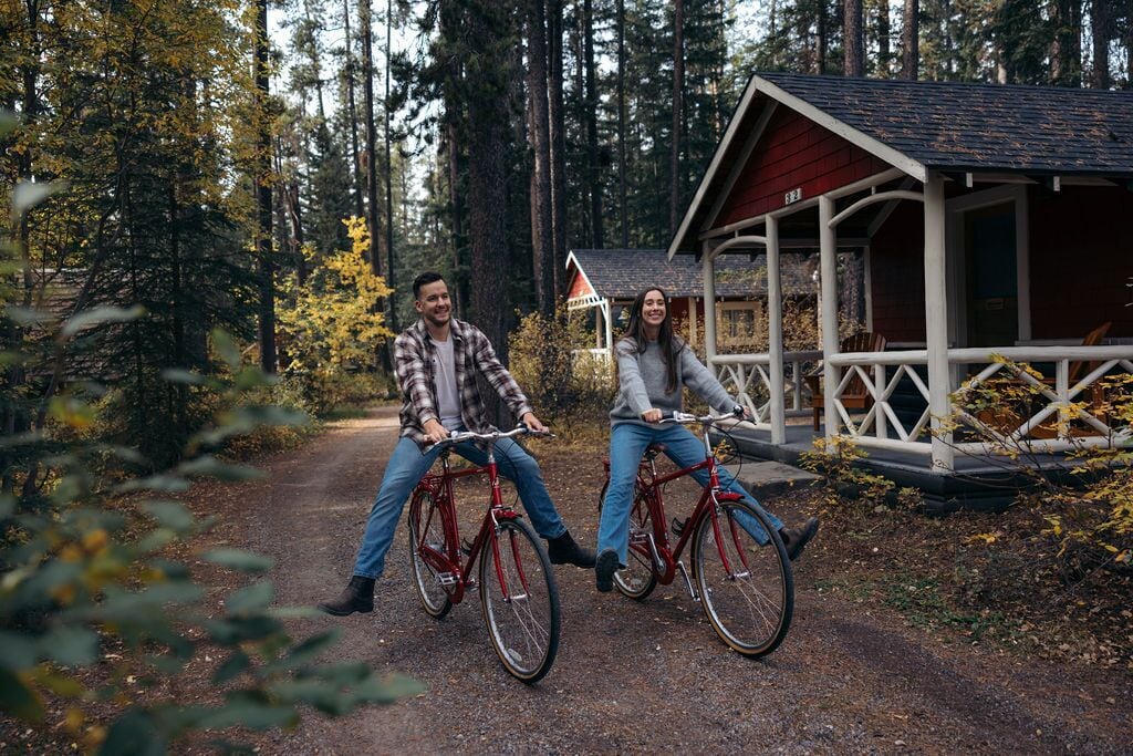 Dos personas en bicicleta sobre un sendero forestal, con una cabaña pequeña al fondo.
