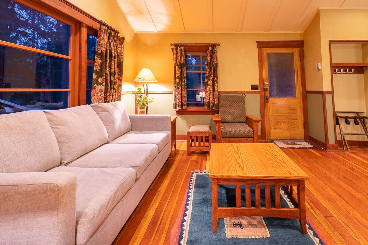 Een gezellige woonkamer met een bank, een houten tafel en een stoel. Er is een raam met gordijnen, een lamp en een deur met kapstok.