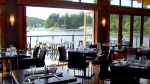 A restaurant with tables set for dining, large windows offering a view of a lake and forest. - Painted Boat Resort (Madeira Park)