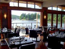 Un restaurante con mesas dispuestas para comer, grandes ventanales que ofrecen vistas a un lago y un bosque.