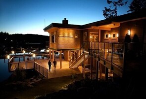 Una cabaña de madera con balcón de cristal, una escalera y una zona de piscina al anochecer.