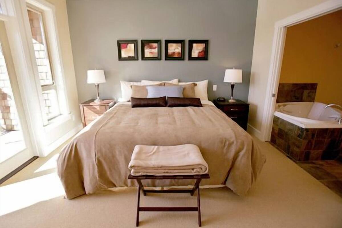 A bedroom with a large bed, bedside tables with lamps, a bench, and a bathroom with a bathtub.