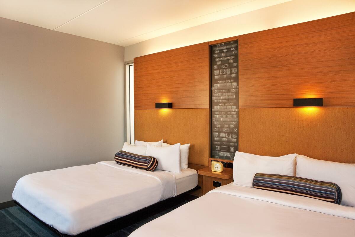 A hotel room with two beds, a wooden headboard, a nightstand with a clock, and a wall-mounted light fixture.