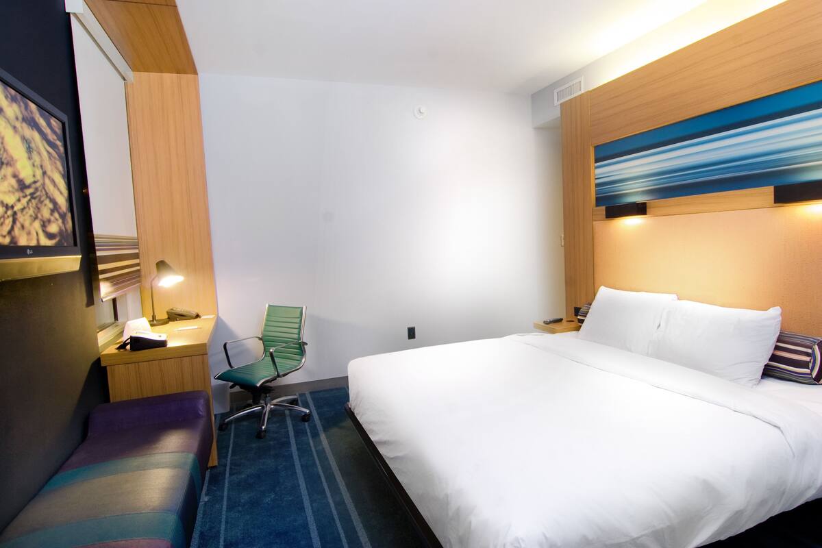A hotel room with a bed, a desk with a chair, a lamp, and a painting on the wall.