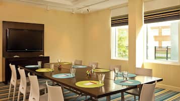 A conference room with a long table, white chairs, a flat-screen TV, and a window with blinds.