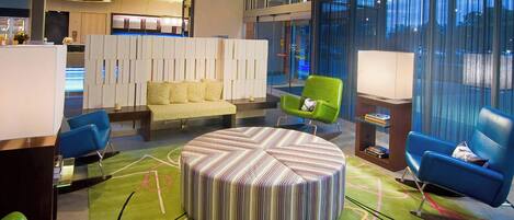 A modern hotel lobby with a round seating area, green and blue chairs, a reception desk, and a large window with curtains.